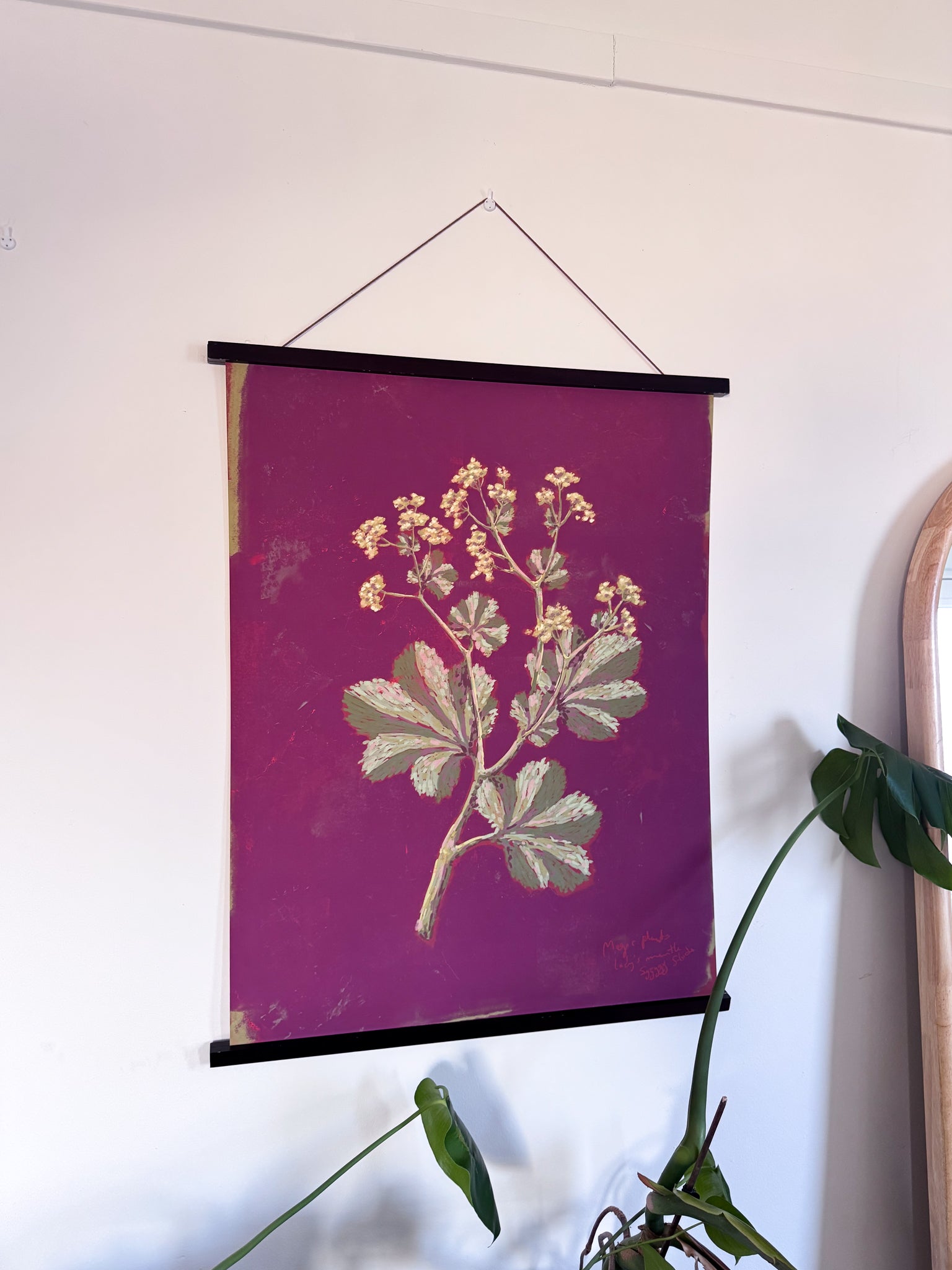 Floral print on a purple background hanging on a wall with a plant in the foreground.