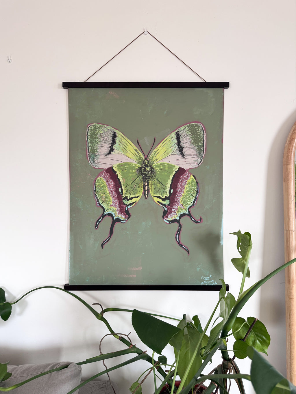 Butterfly illustration on a green background hanging on a wall with plants in the foreground.
