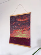 Artistic depiction of a sunset with vibrant colors on a wooden hanging frame against a white wall.