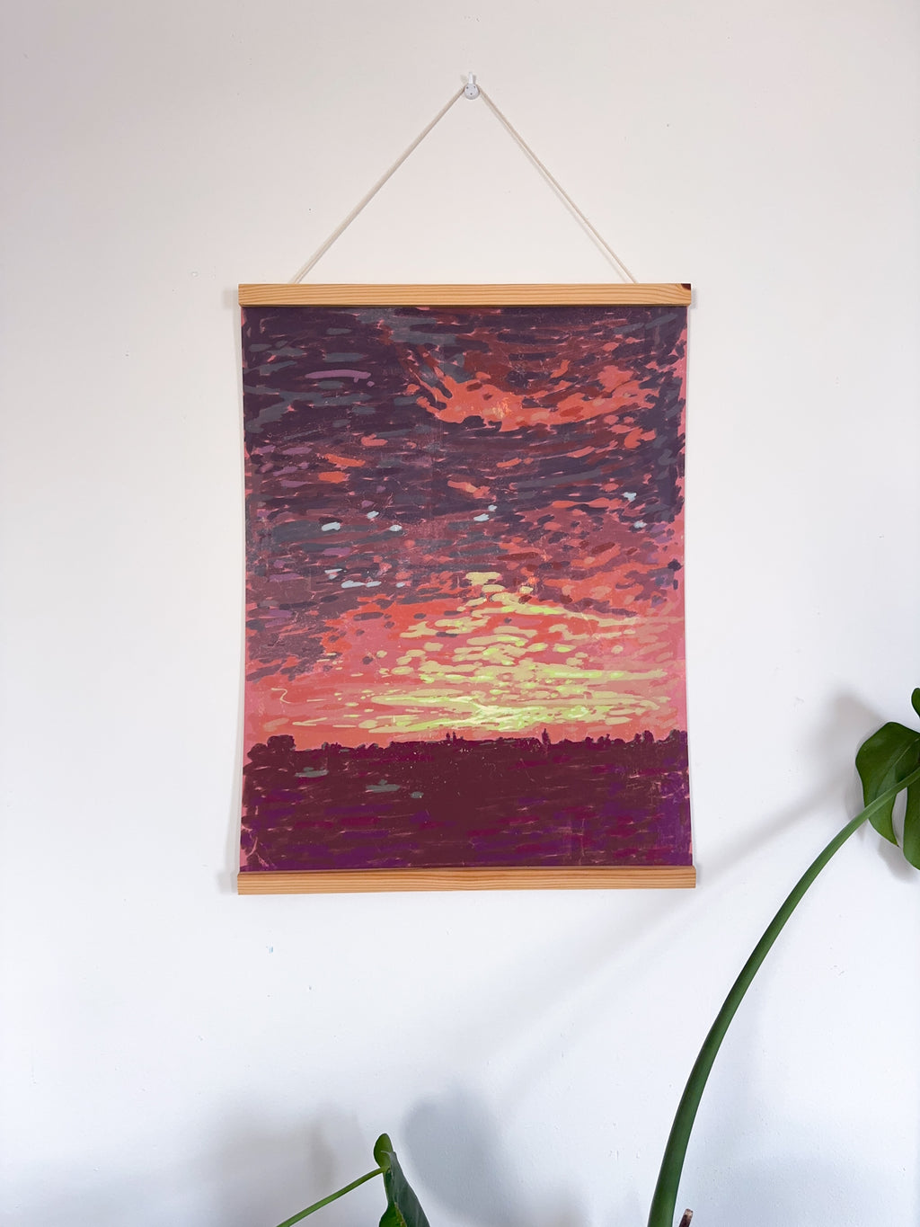 Artwork of a sunset landscape on a white wall with a plant in the corner.