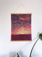 Artwork of a sunset landscape on a white wall with a plant in the corner.