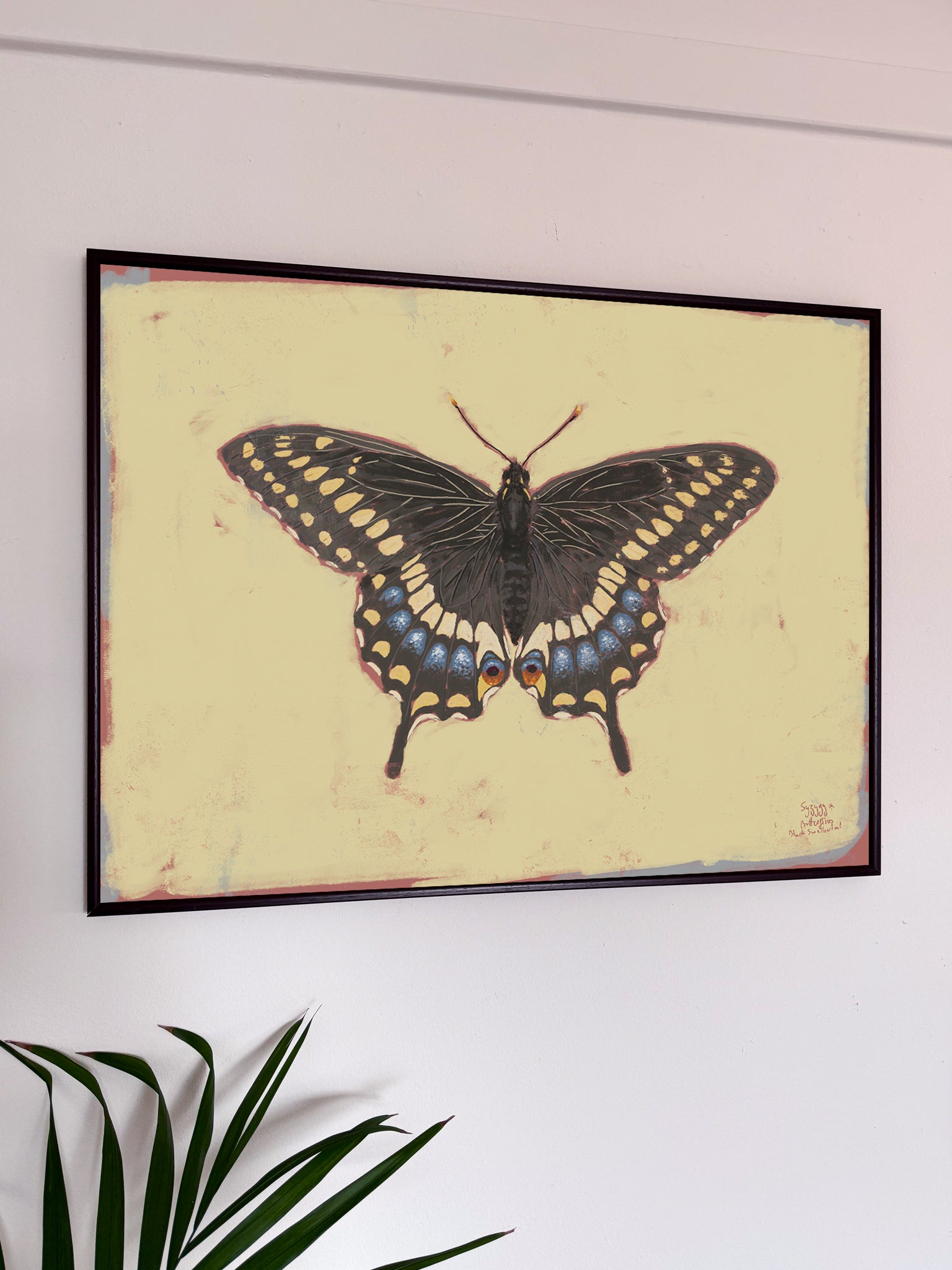 Framed butterfly artwork on a white wall with a plant in the foreground.