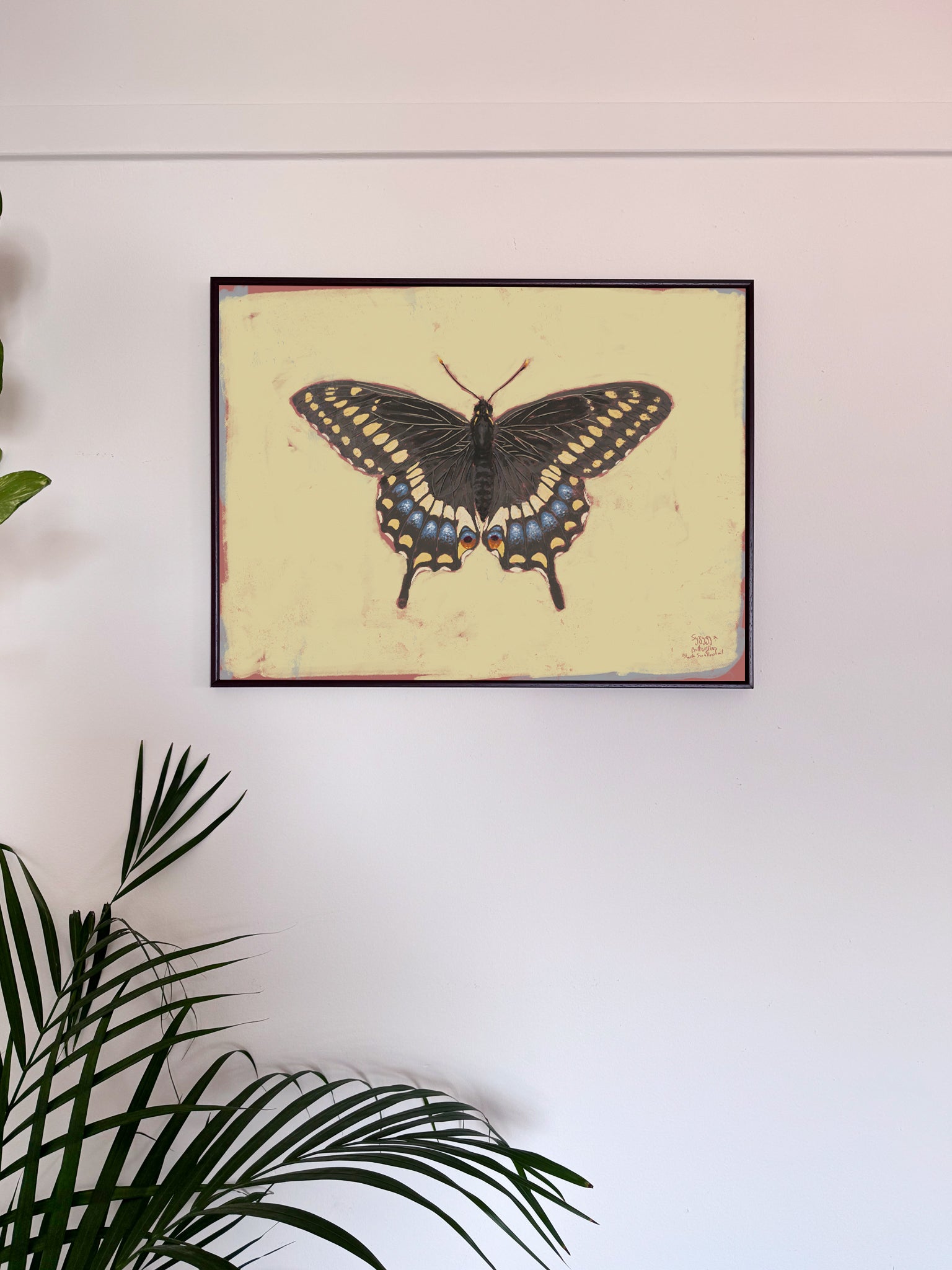 Framed butterfly artwork on a white wall with green leaves in the foreground