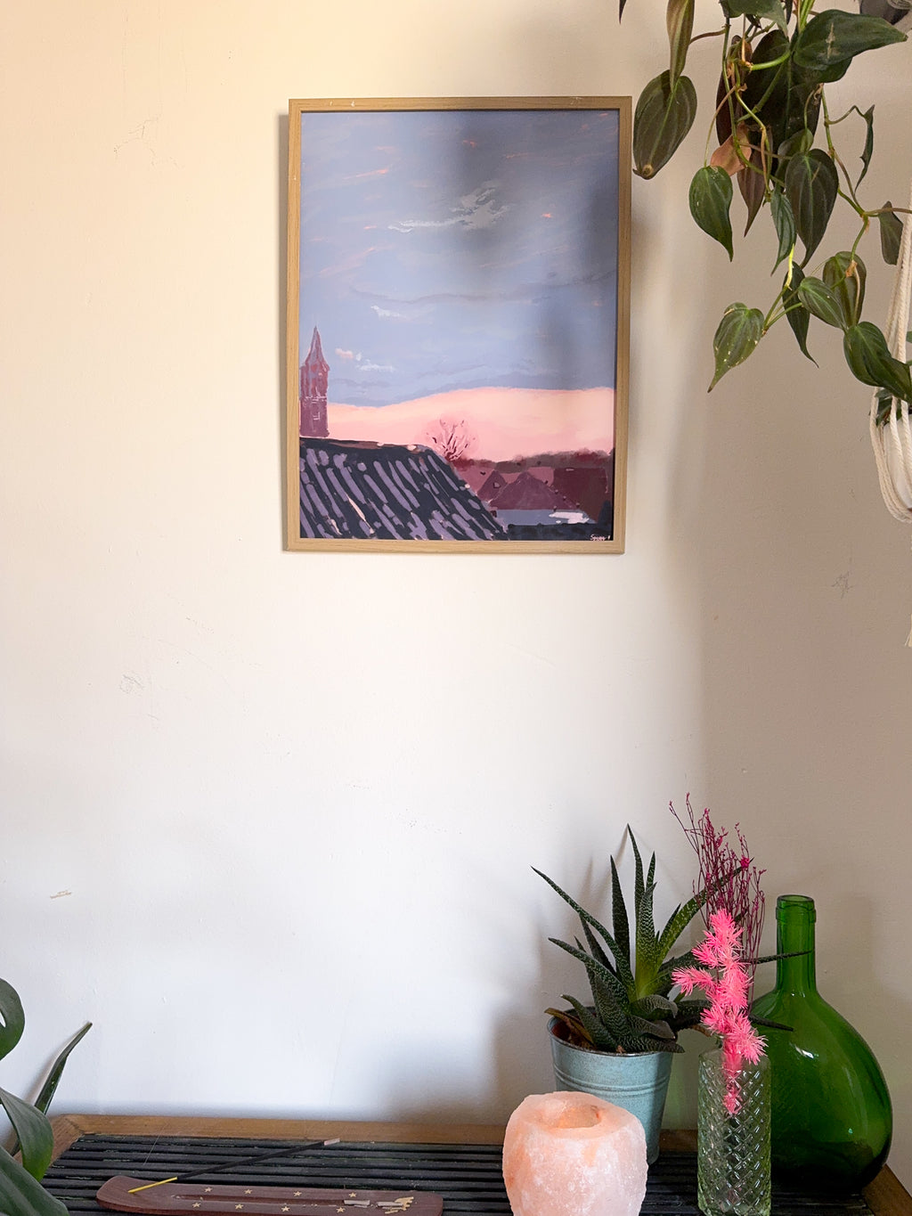 Framed artwork of a city landscape and pink cloud, on a wall with plants and decorative items below