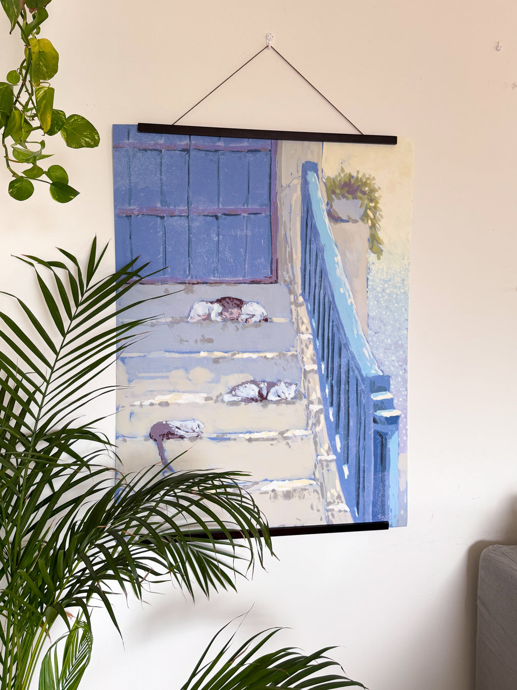Poster of a blue door and cats sleeping on the staircase, on a wall with a plant in the foreground