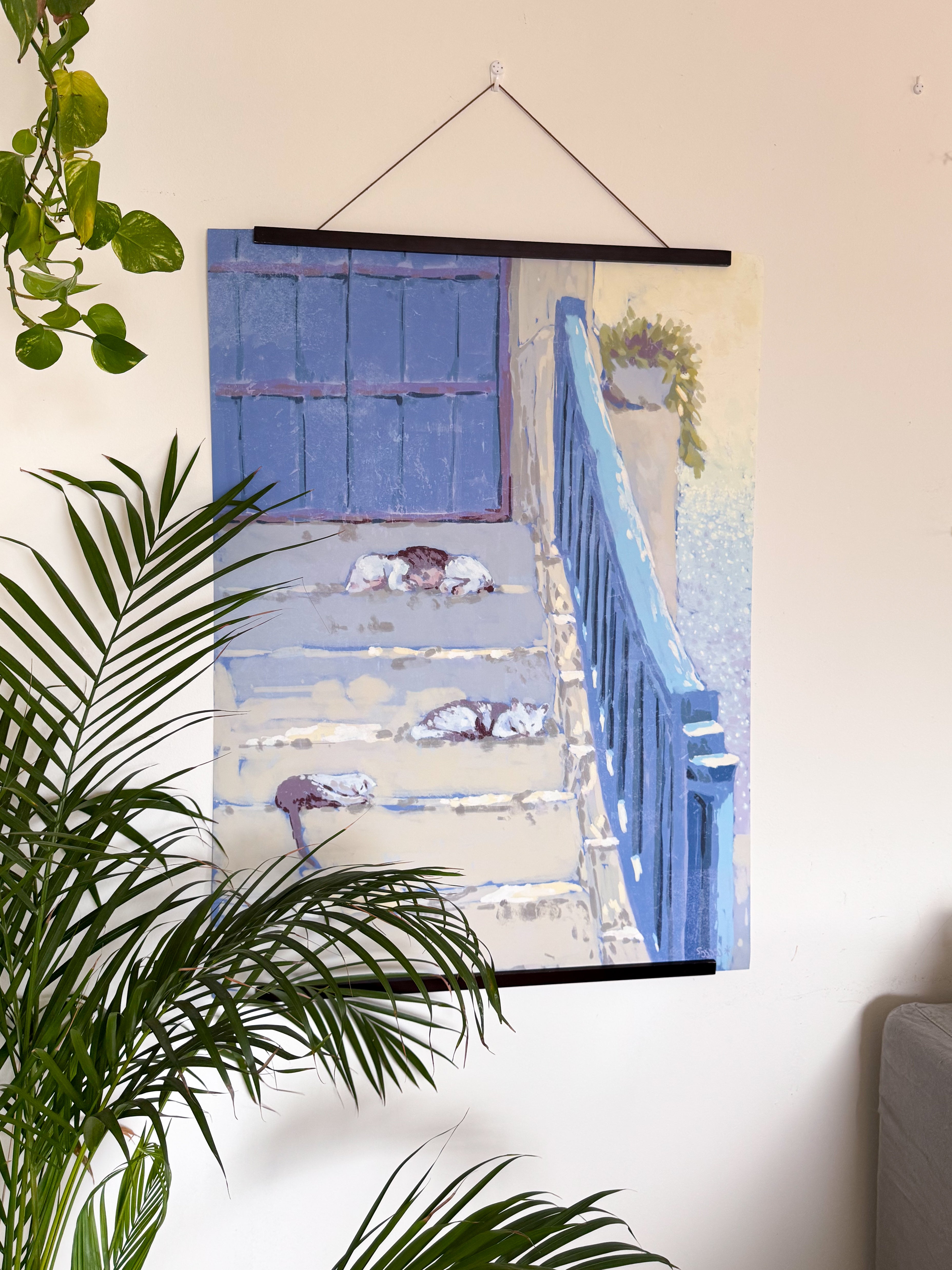 Poster of a blue door and cats sleeping on the staircase, on a wall with a plant in the foreground