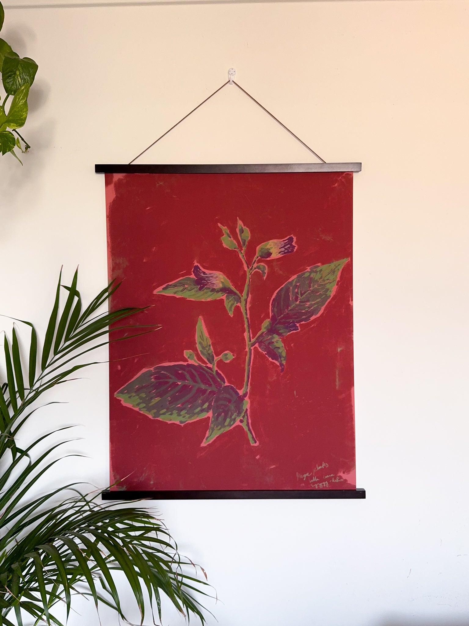 Decorative art print with floral design on a red background, hanging against a white wall with green plants.