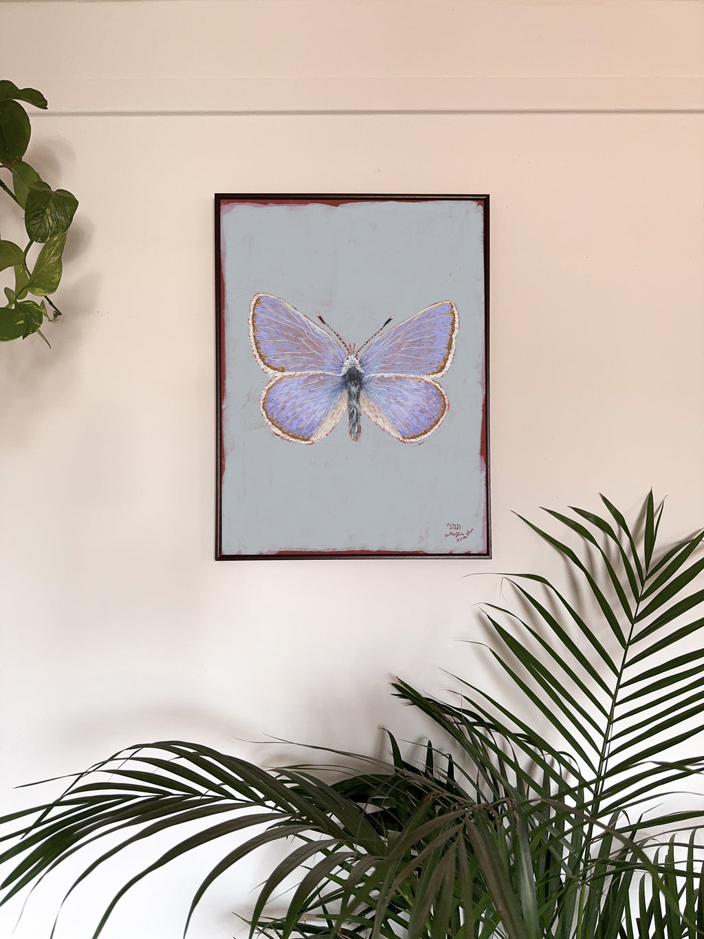 Framed artwork of a butterfly on a light blue background, surrounded by green plants.