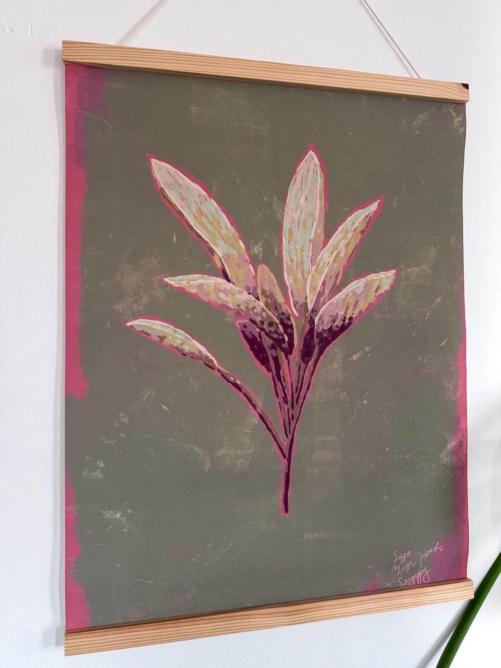 Floral sage artwork with pink leaves in a wooden magnetic frame hanging on a white wall, side angle.