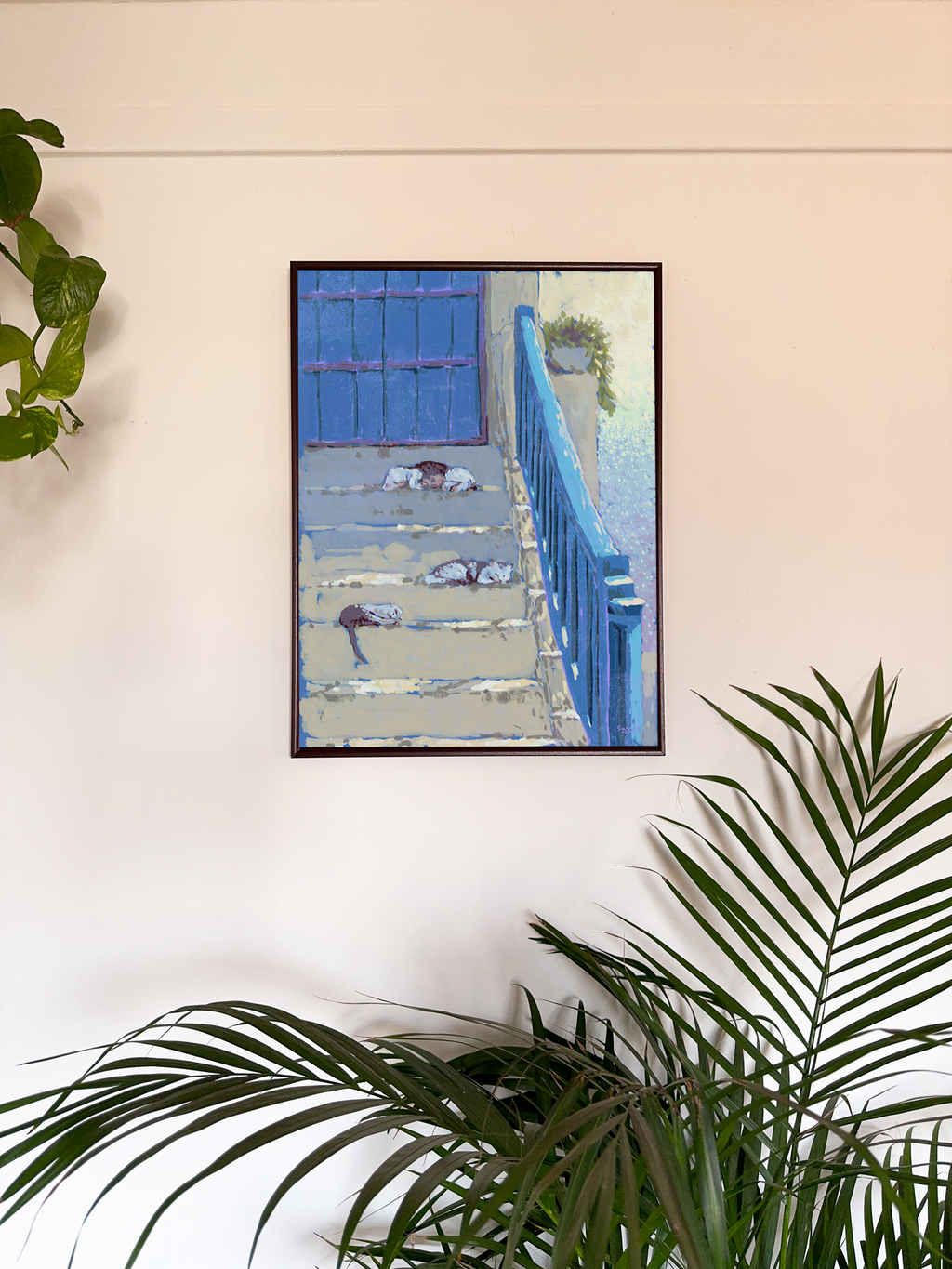 Framed artwork of cats lying on a staircase with the sea in background displayed on a wall and a plant